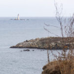 A white lighthouse is seen in the distance from a rocky ocean shoreline